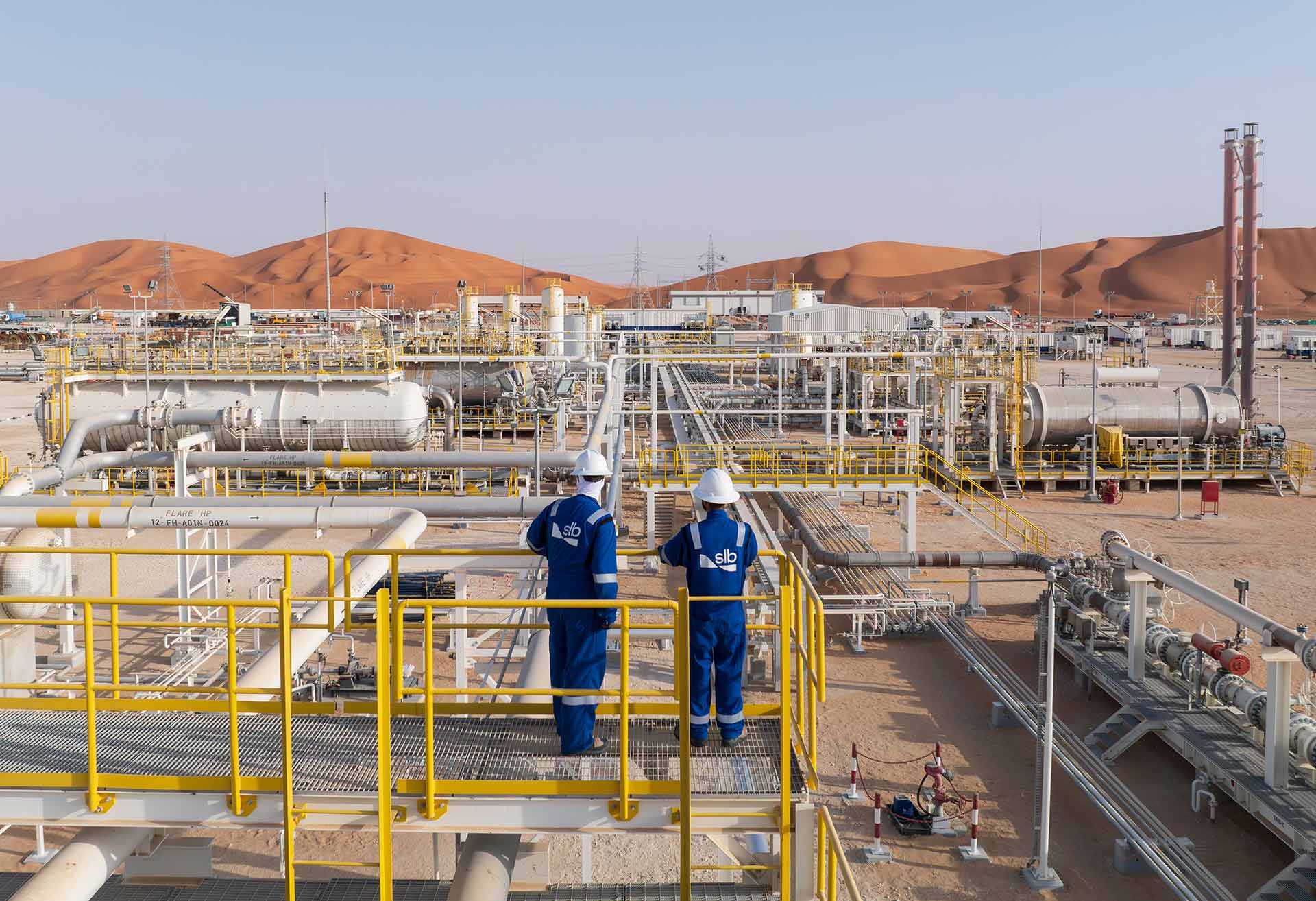Photo of 2 SLB employees in coveralls and hard hats looking across a processing facility in a desert.
