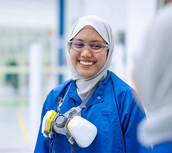 SLB team member in a blue lab coat and safety gear, smiling in a high-tech facility—representing diversity, safety, and innovation in the workplace.
