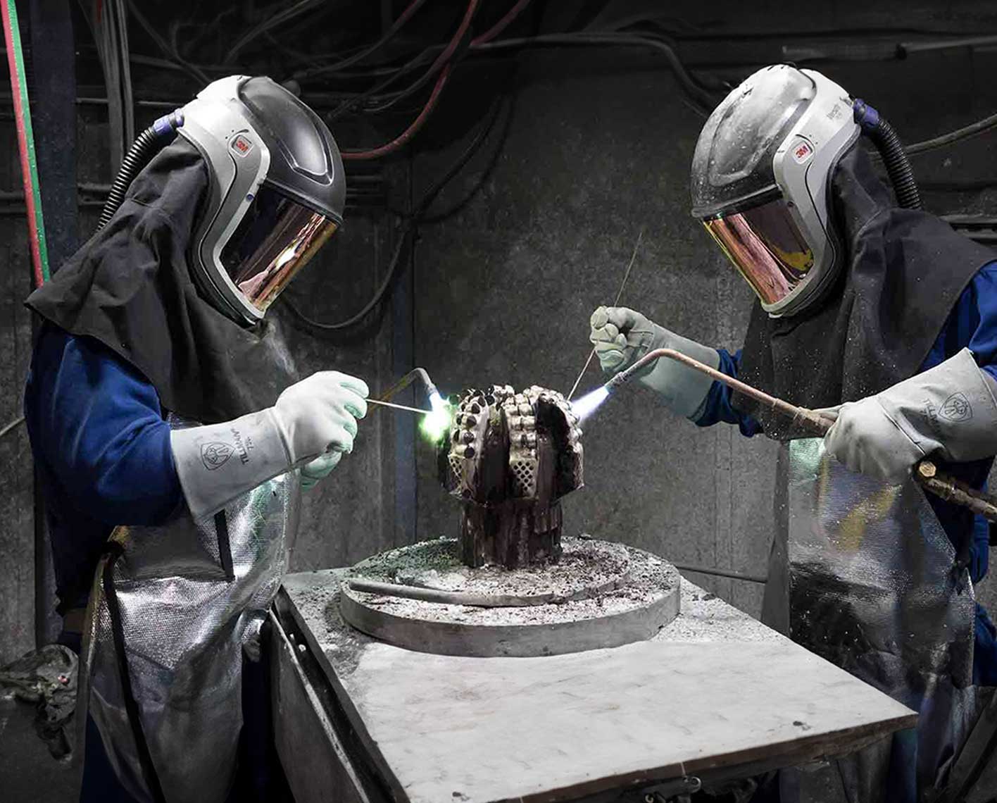 Two people in PPE welding a drill bit.