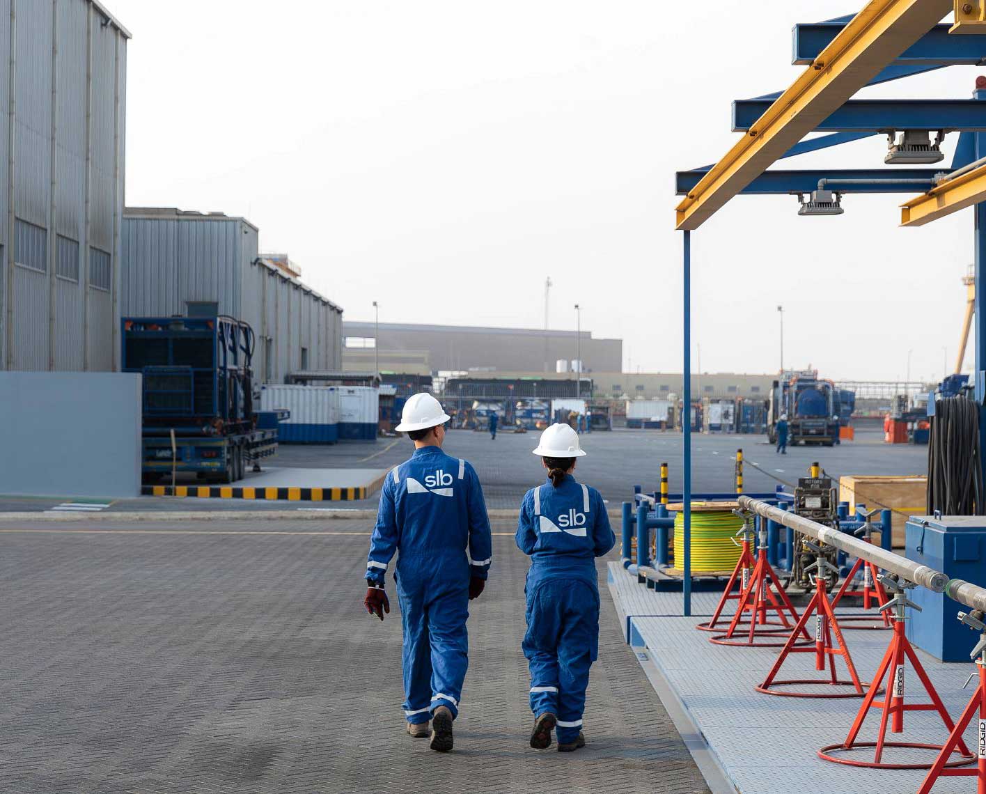 Two workers in blue coveralls and white hard hats walking through an industrial yard with equipment