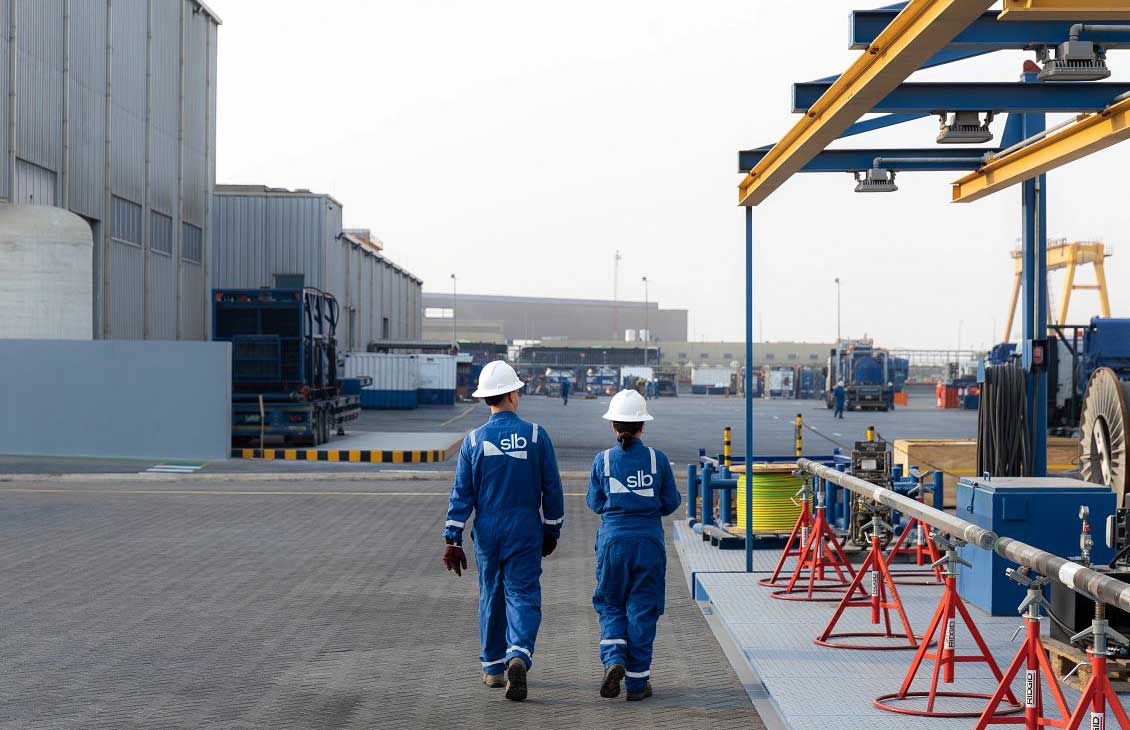 Two workers in blue coveralls and white hard hats walking through an industrial yard with equipment