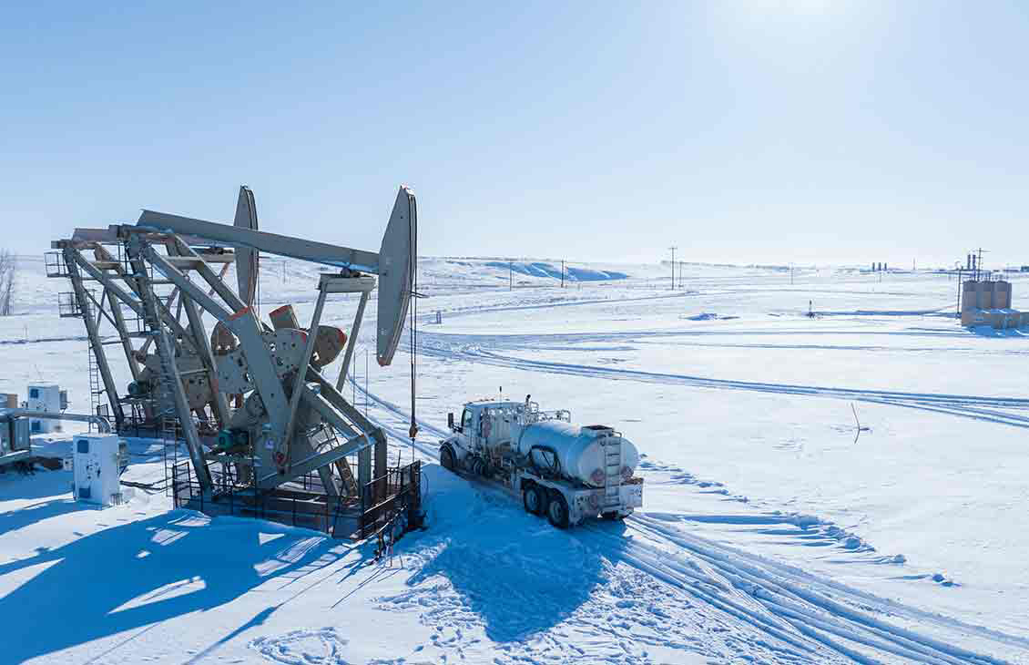 A pumpjack, oil tanker, and storage tanks in a snow-covered oil field.