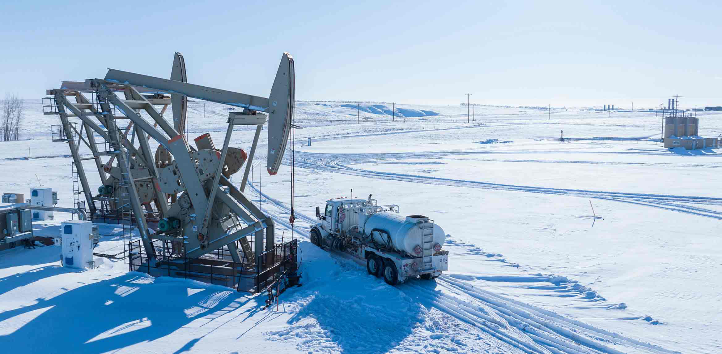 A pumpjack, oil tanker, and storage tanks in a snow-covered oil field.