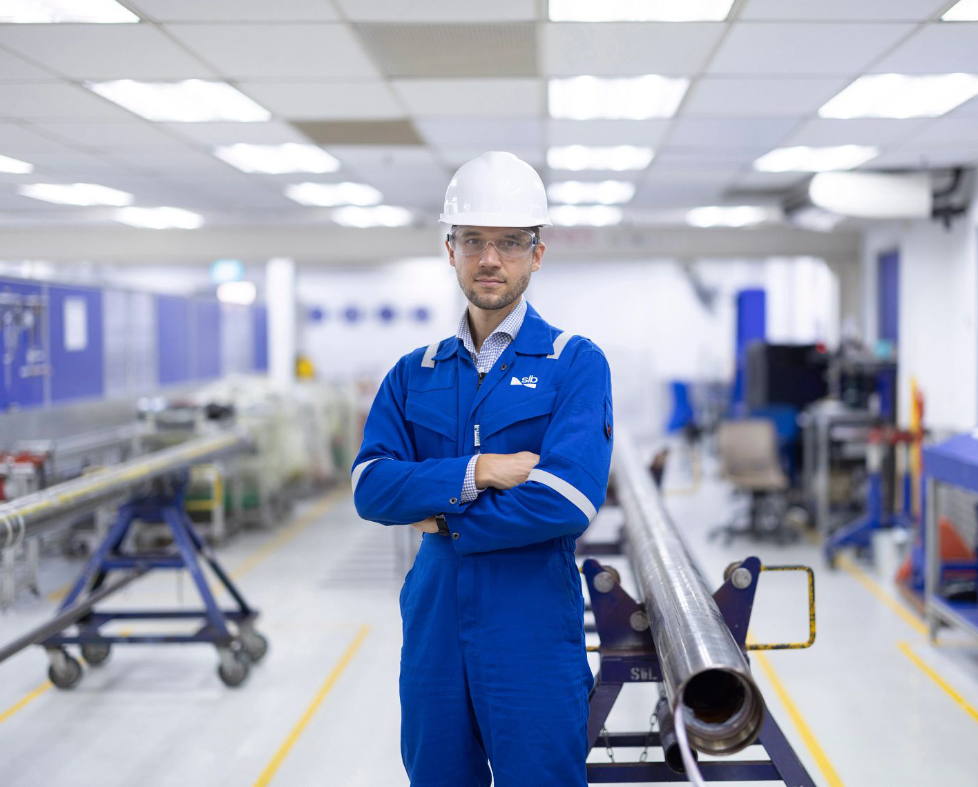 Engineer wearing blue coveralls, a white hard hat, and safety glasses standing with arms crossed in an industrial testing facility beside a section of pipe.