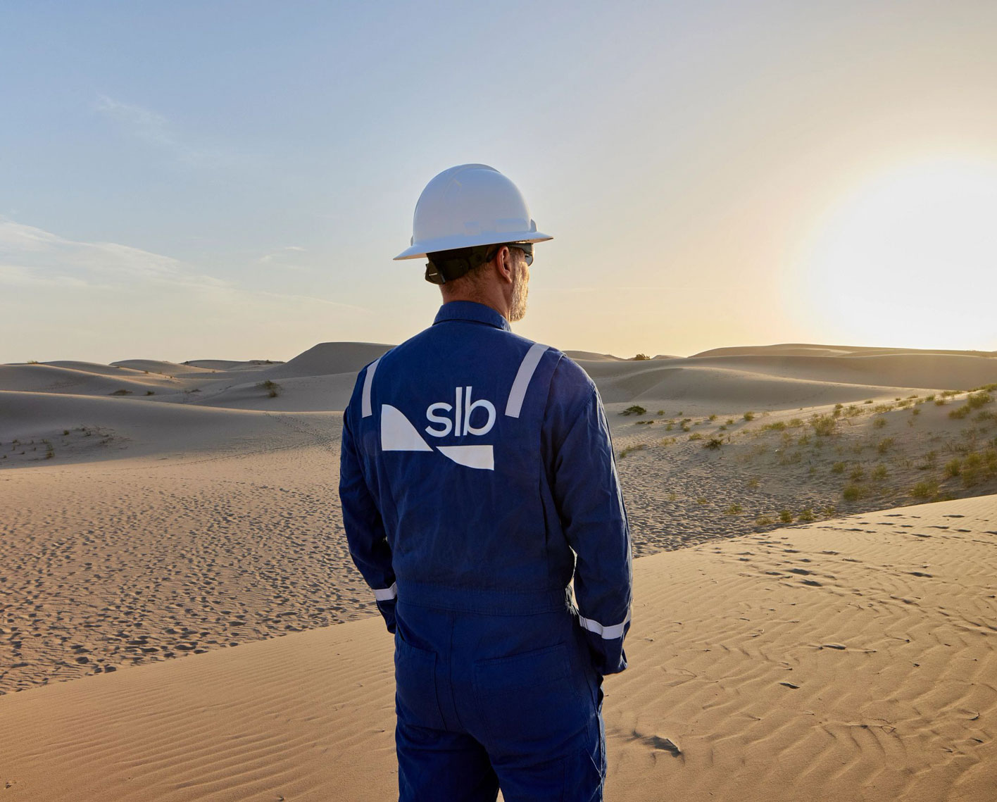 A man in a white hard hat and blue coveralls with "SLB" printed on the back looking into a field