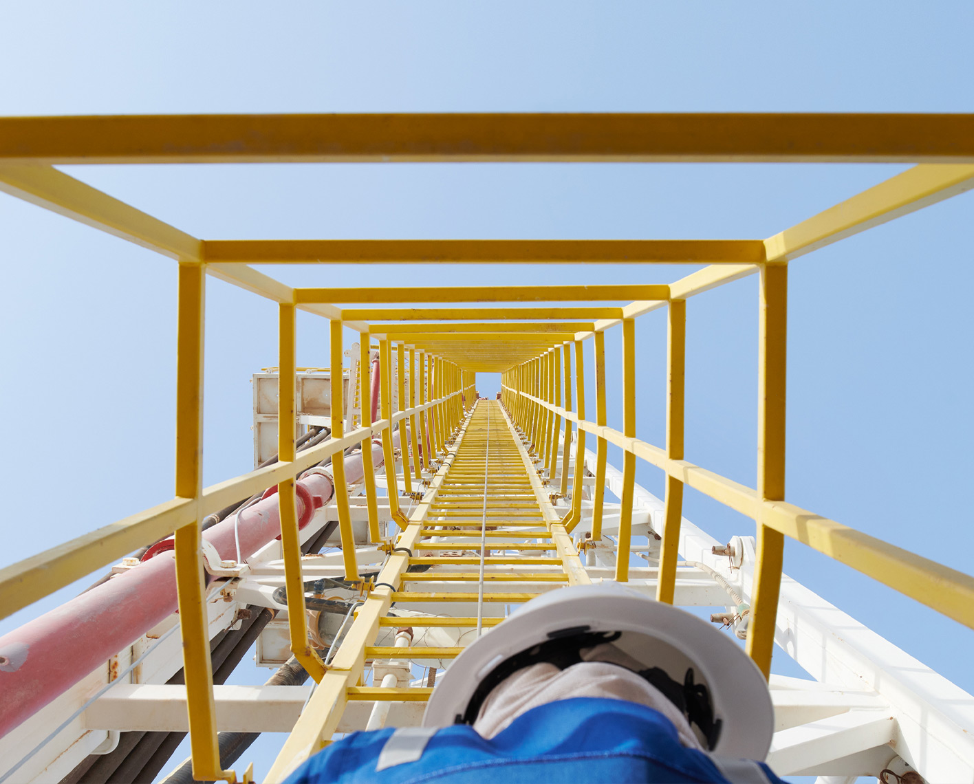 Upward view of a man in blue coveralls and white helmet looking up at a tall ladder