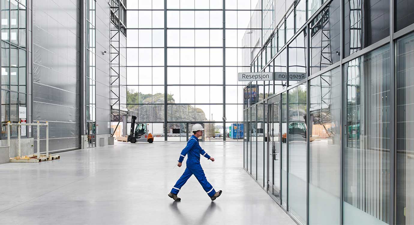 man in blue coveralls walking across a warehouse with large windows in the background.
