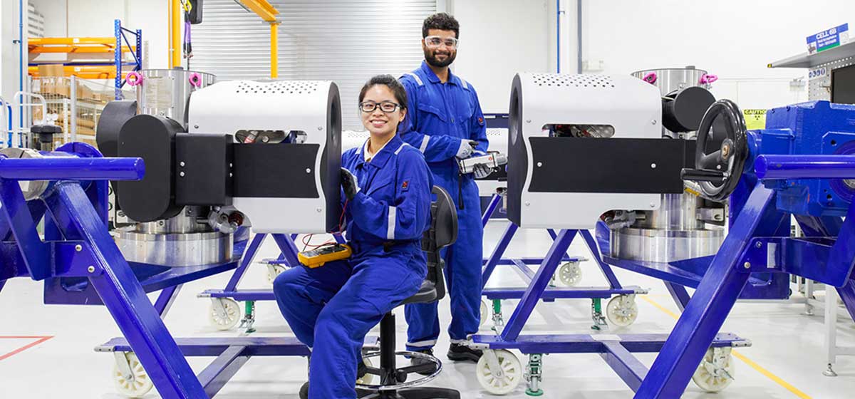 2 employees in blue coveralls and safety glasses smiling while working on a Vx Spectra