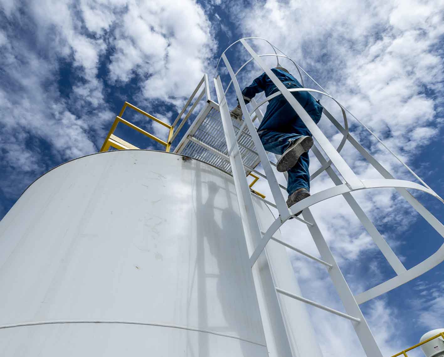 a man climbing a ladder on a storage tank, shot from below looking up towards the sky with clouds