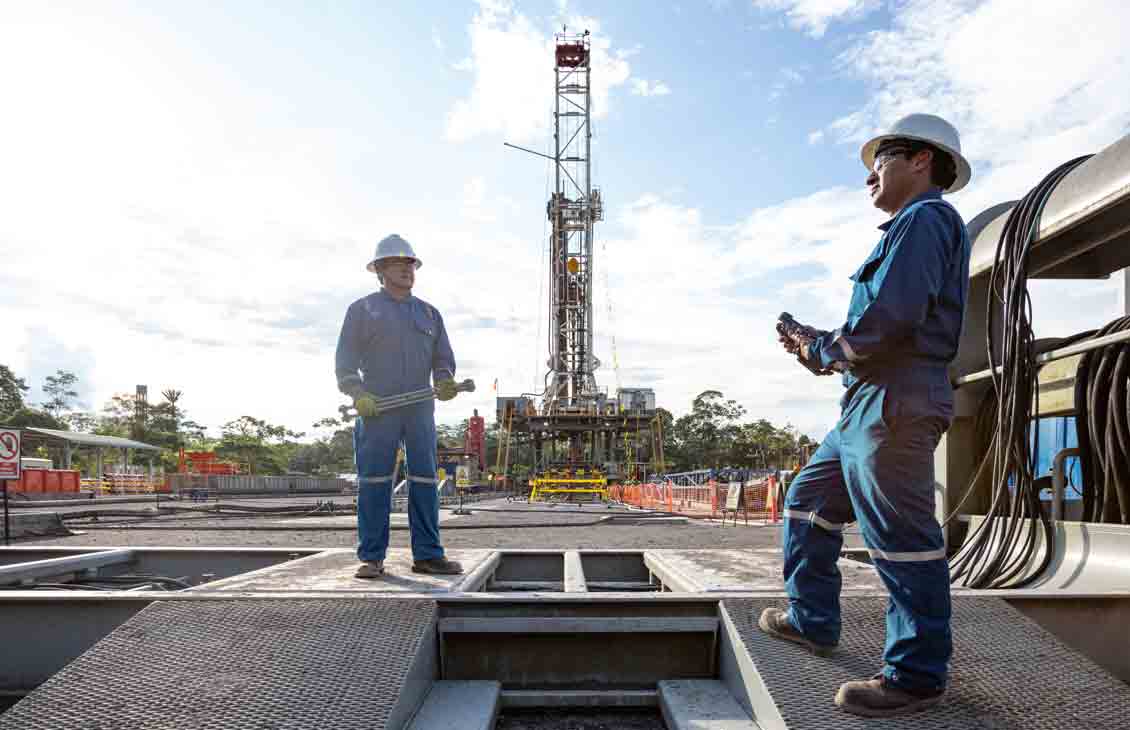Photo of men in blue coveralls working on rig site - rig in background 