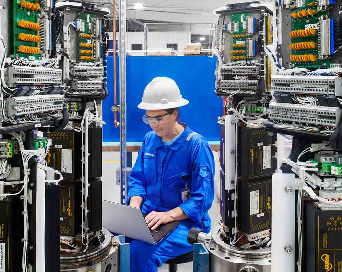 Woman in SLB blue coveralls working on a laptop among shelfs of wires and computers