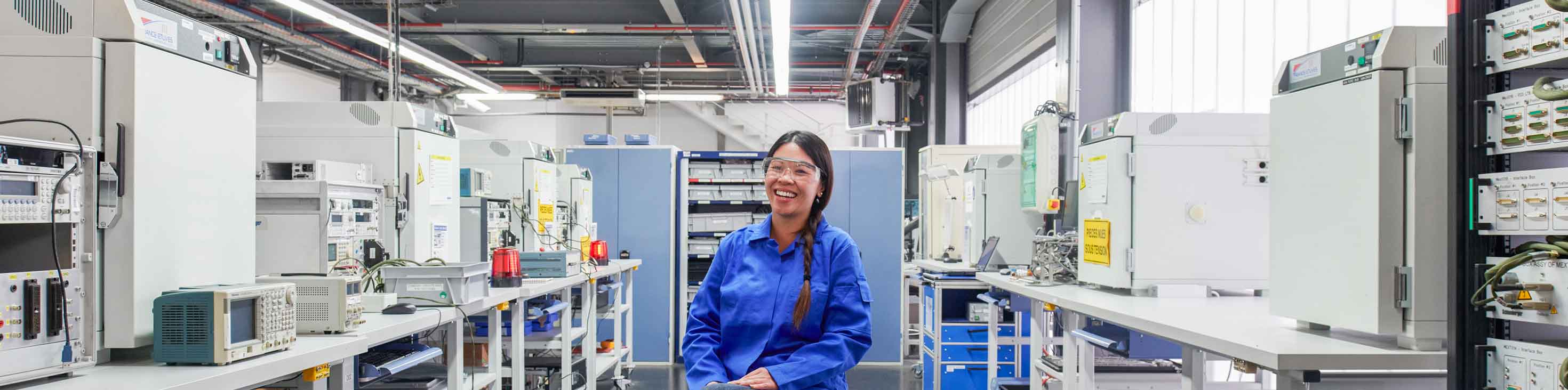 Woman in safety goggles, black pants, and blue lab coat sitting on still smiling in lab