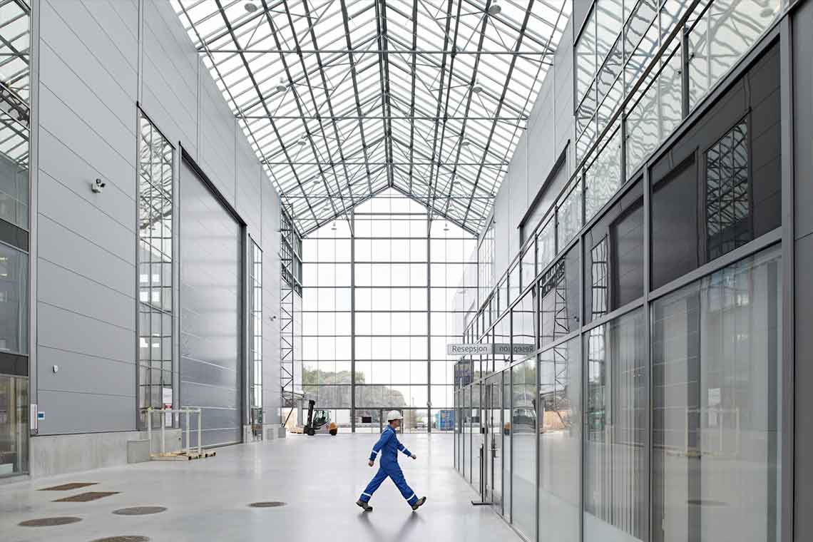 Man in blue coveralls and white hardhat walking in warehouse with glass roof