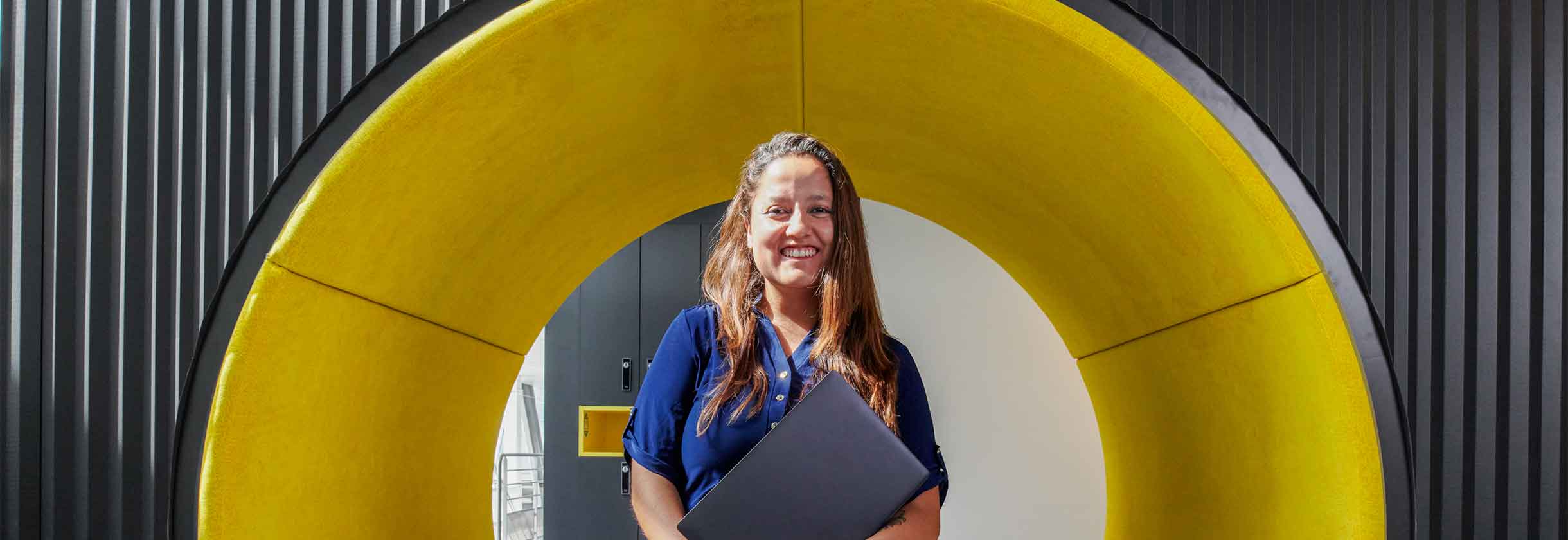 woman in khaki pants and blue polo smiling standing in front of yellow arch with benches