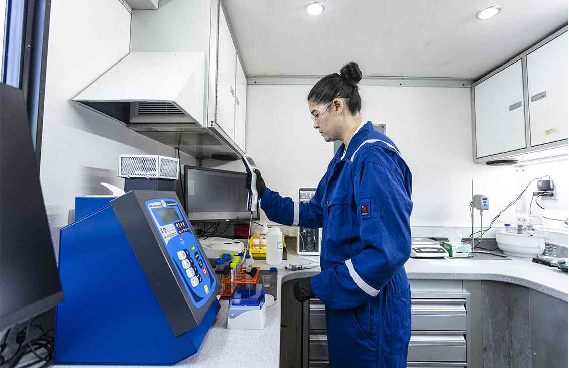 Woman in lab wearing blue coveralls using probe on test tubes full of liquid