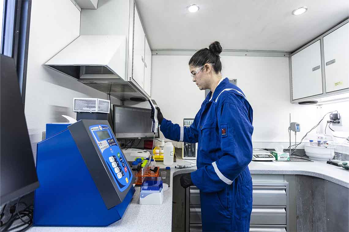 Woman in lab wearing blue coveralls using probe on test tubes full of liquid