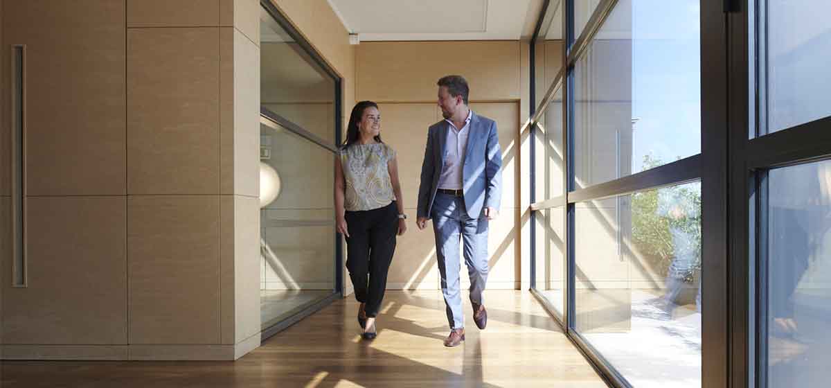 Man in blue suit and woman in white shirt and blue slacks walking down an office hallway, with windows on one side.