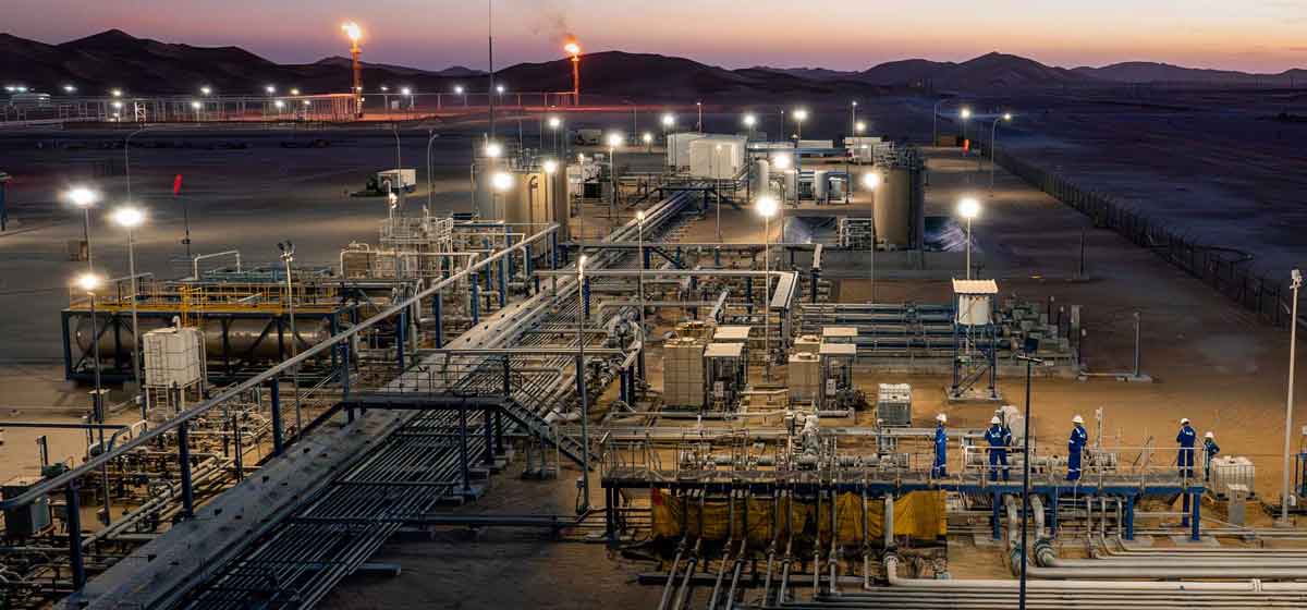 Industrial oil and gas facility at dusk with workers and lit flare stacks in a desert setting.
