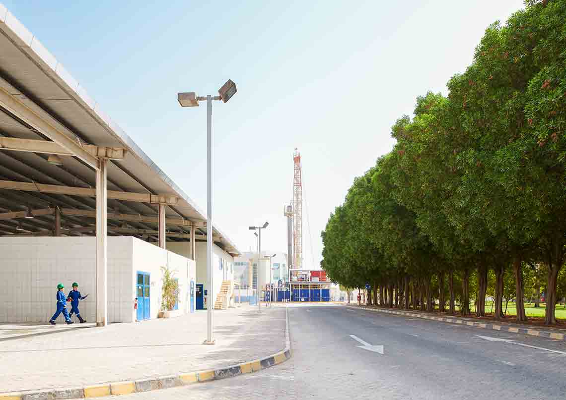 Landscape photo of buildings on a street, rig in the background, and people in blue coveralls walking