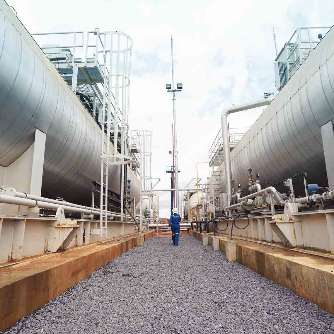 Photo of person in blue coveralls walking between large tanks and pipes