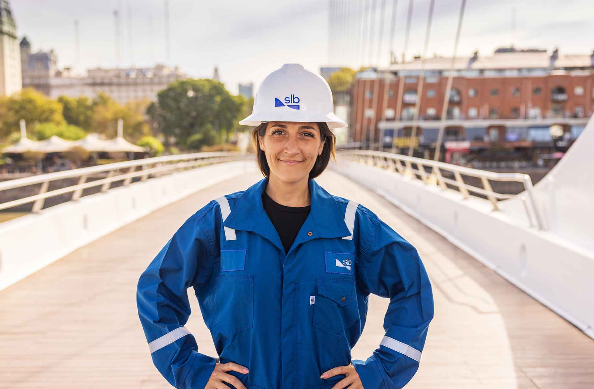 Female SLB employee in blue coveralls standing on a bridge with a city in the background