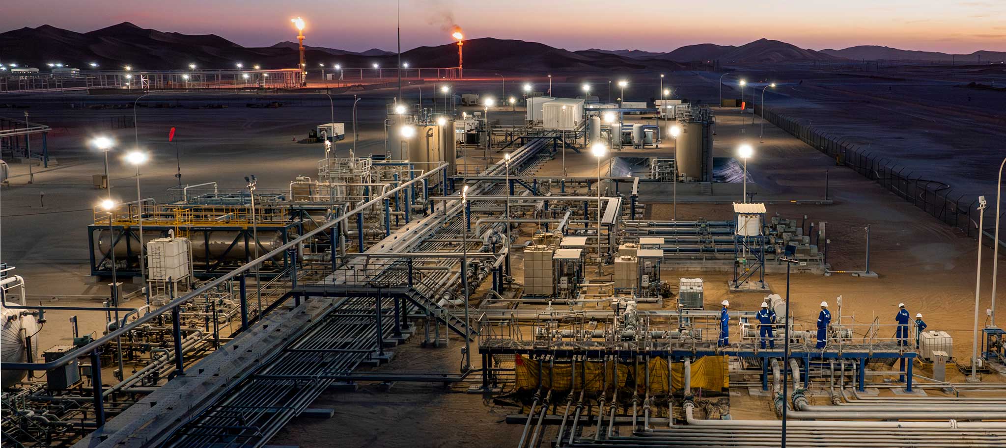 Industrial oil and gas facility at dusk with workers and lit flare stacks in a desert setting.