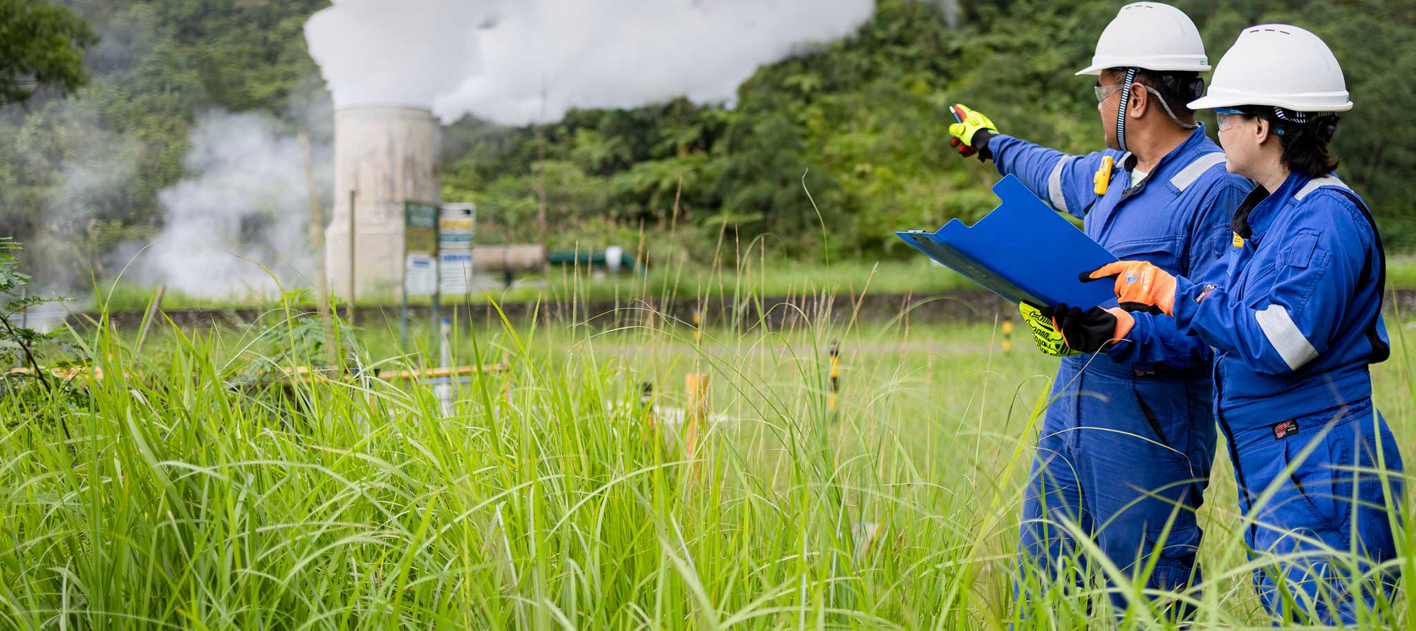 Two workers in safety gear point toward a steaming vent in a grassy area.