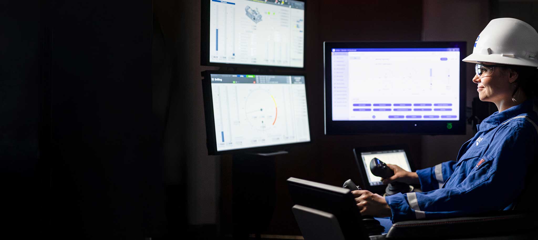 A woman in safety gear, including a hard hat and protective glasses, operates an advanced control system in a dark room with multiple digital screens displaying technical data and drilling information.