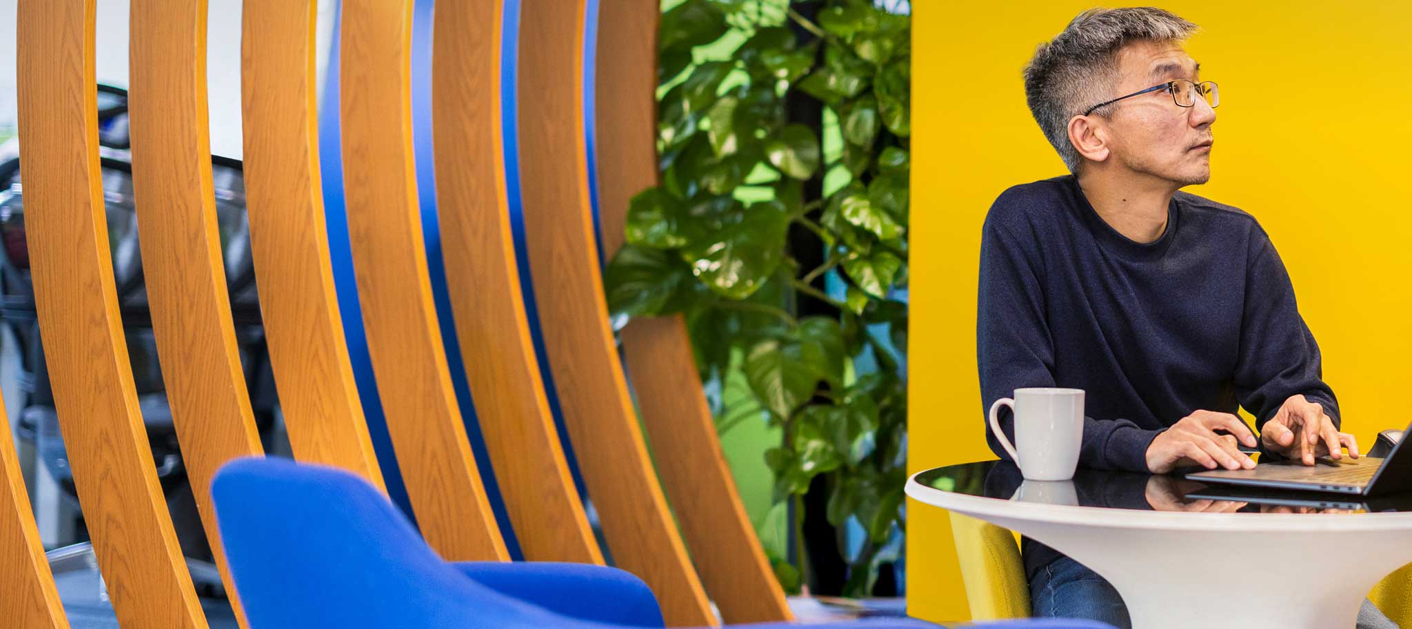 Man using a laptop at a small table with a mug, against a bright yellow wall in a modern office.