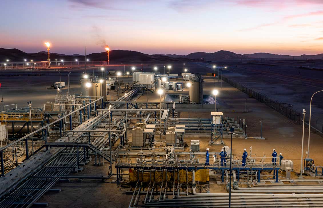 Industrial oil and gas facility at dusk with workers and lit flare stacks in a desert setting.