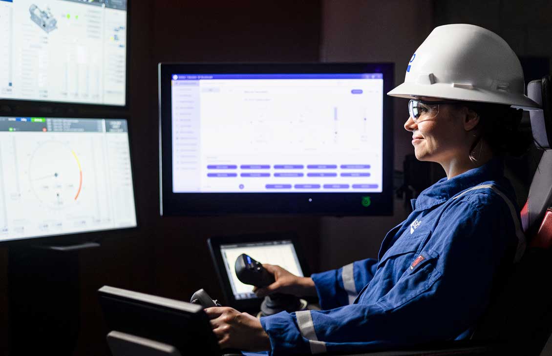 A woman in safety gear, including a hard hat and protective glasses, operates an advanced control system in a dark room with multiple digital screens displaying technical data and drilling information.