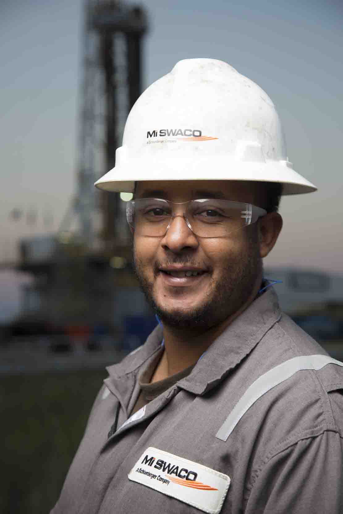 Person in coveralls and hard hat standing in front of drilling rig.