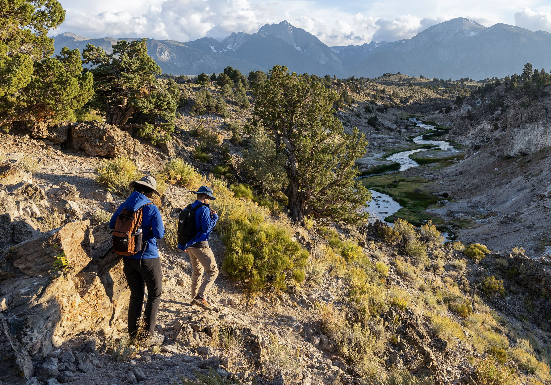 Two people hiking outdoors in rocky terrain by stream and mountains.