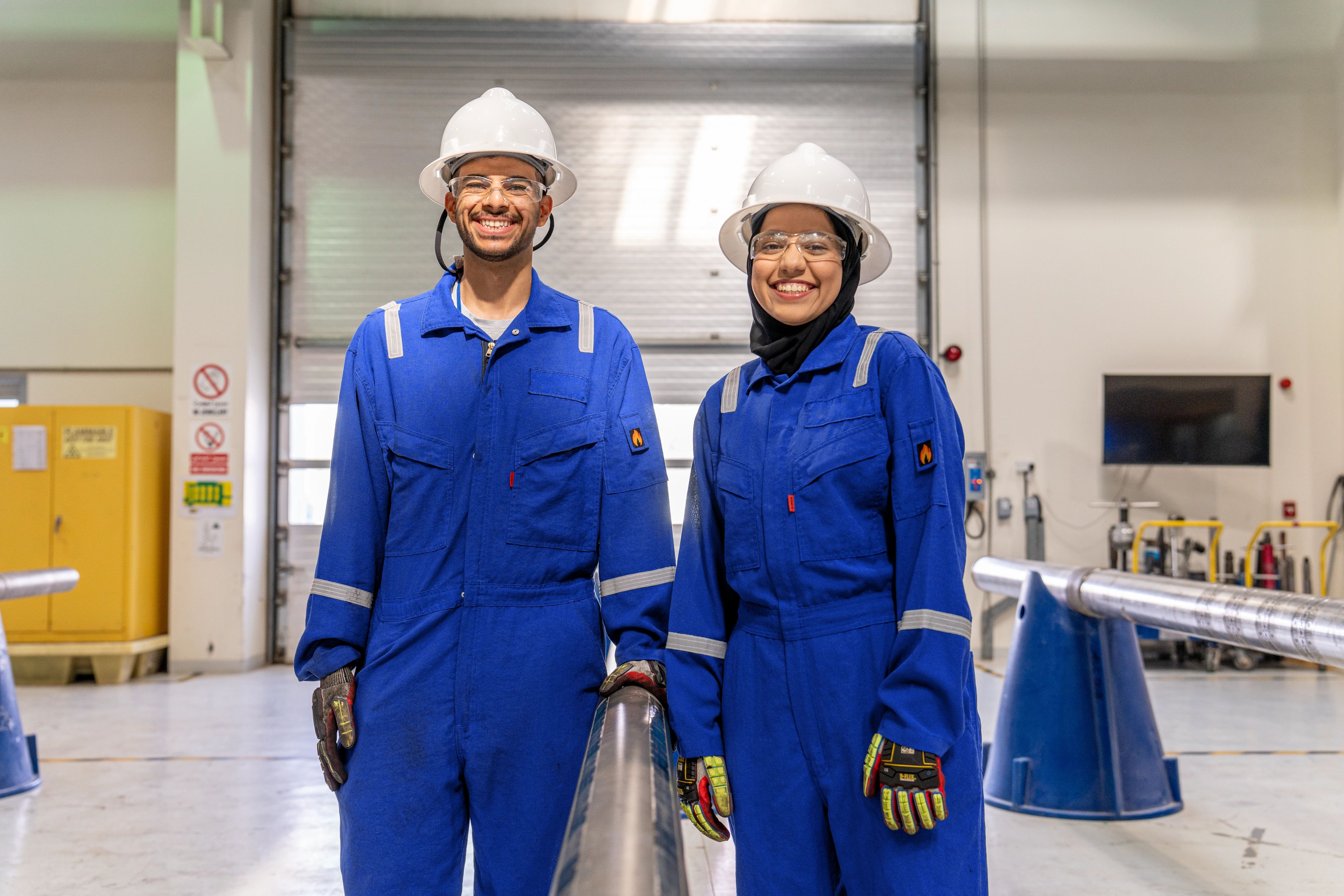 Two SLB employees in blue coveralls smiling with white hard hats on.