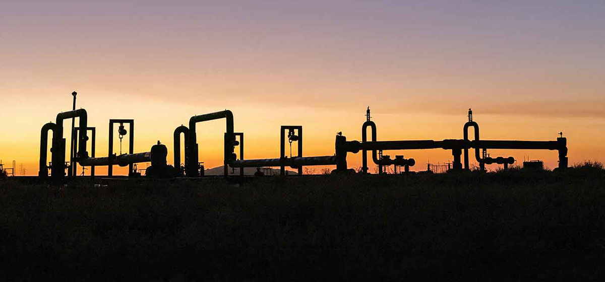 Photo of geothermal wellheads and pipelines silhouetted against an orange sky.